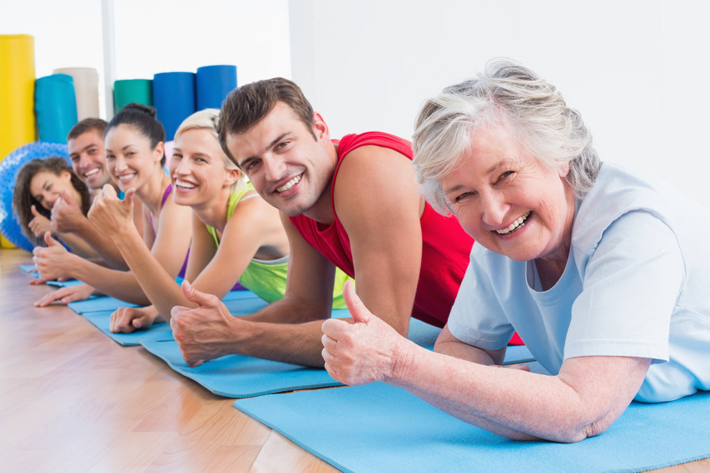 Portrait of happy senior woman with friends gesturing thumbs up while lying on exercise mats at gym
